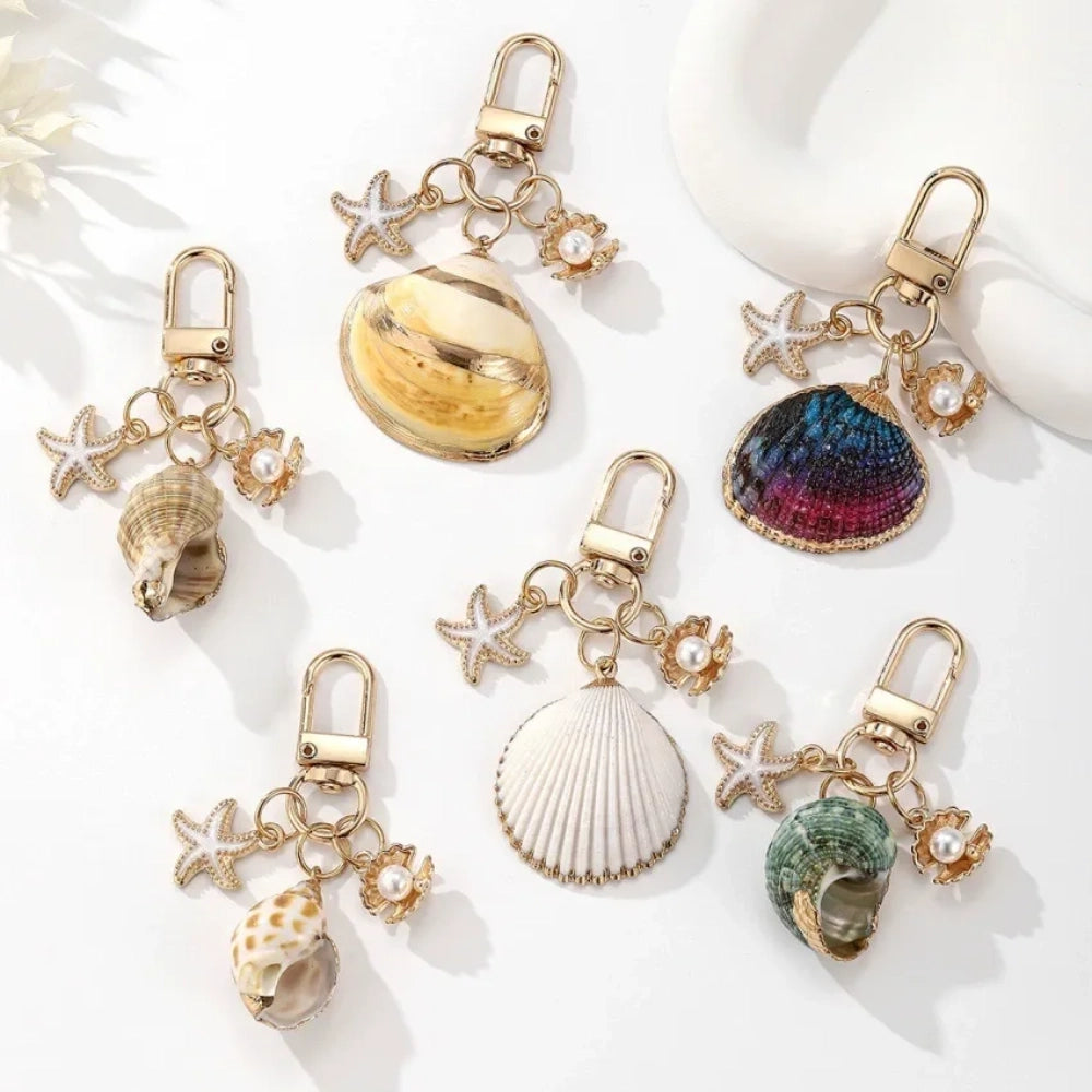 Five ocean-themed keychain charms featuring real shells including conch, scallop, clam, and abalone styles with gold-tone starfish and pearl crab accent charms on swivel clasps.