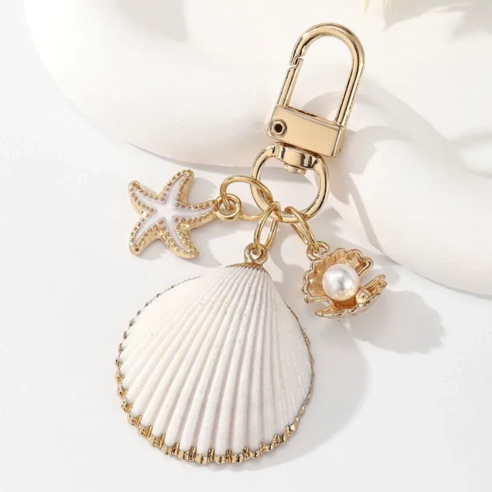 Five ocean-themed keychain charms featuring real shells including conch, scallop, clam, and abalone styles with gold-tone starfish and pearl crab accent charms on swivel clasps. - 1