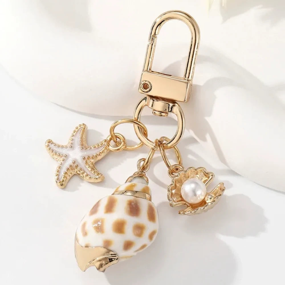Five ocean-themed keychain charms featuring real shells including conch, scallop, clam, and abalone styles with gold-tone starfish and pearl crab accent charms on swivel clasps. - 2