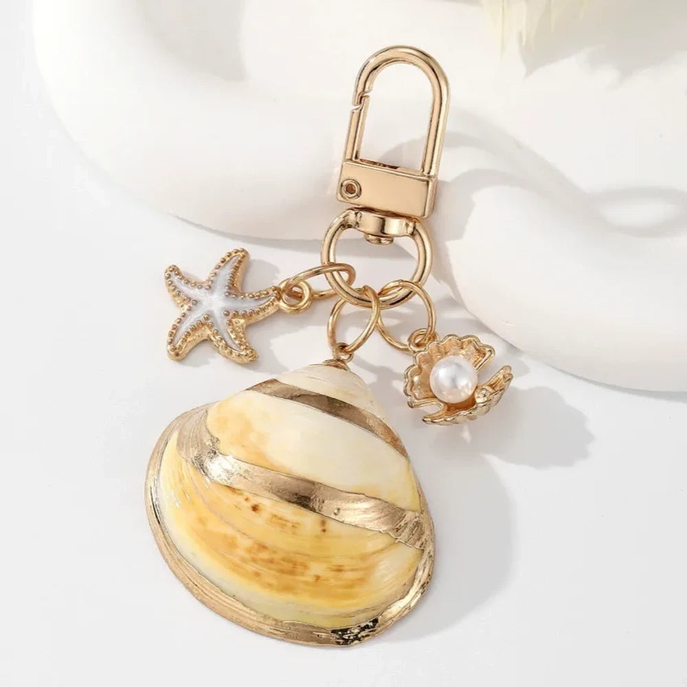Five ocean-themed keychain charms featuring real shells including conch, scallop, clam, and abalone styles with gold-tone starfish and pearl crab accent charms on swivel clasps. - 3