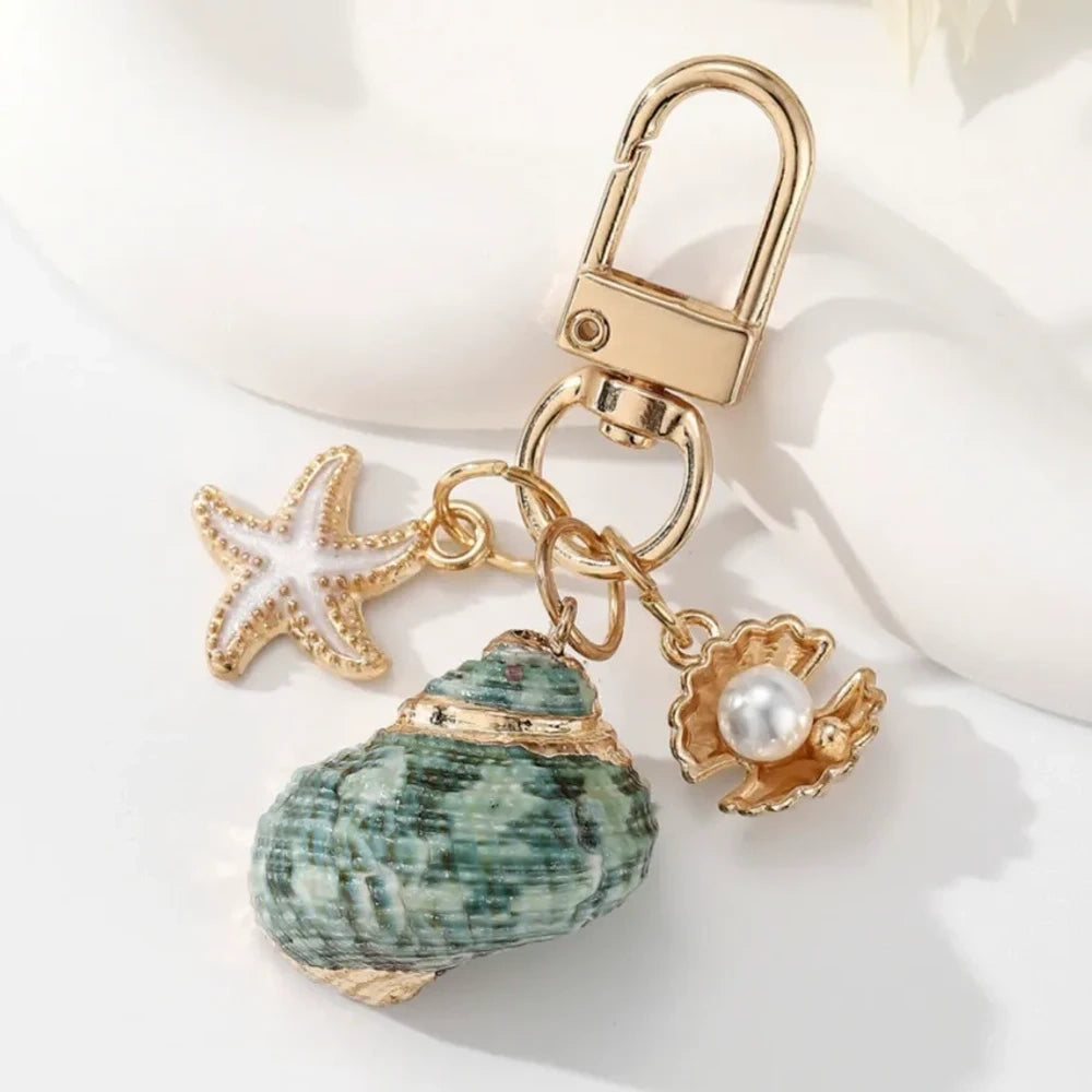 Five ocean-themed keychain charms featuring real shells including conch, scallop, clam, and abalone styles with gold-tone starfish and pearl crab accent charms on swivel clasps. - 4
