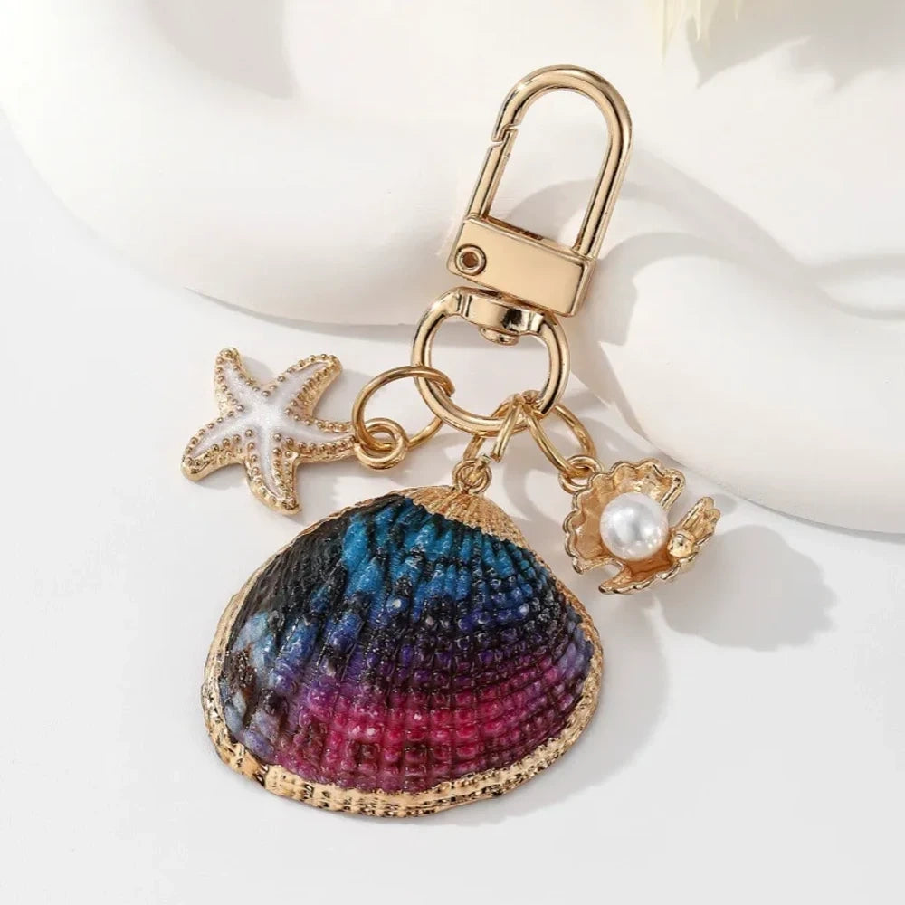 Five ocean-themed keychain charms featuring real shells including conch, scallop, clam, and abalone styles with gold-tone starfish and pearl crab accent charms on swivel clasps. - 5