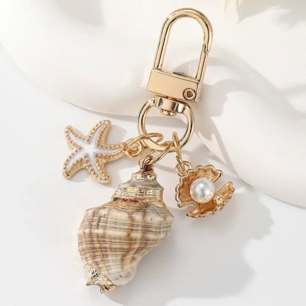 Five ocean-themed keychain charms featuring real shells including conch, scallop, clam, and abalone styles with gold-tone starfish and pearl crab accent charms on swivel clasps. - 6