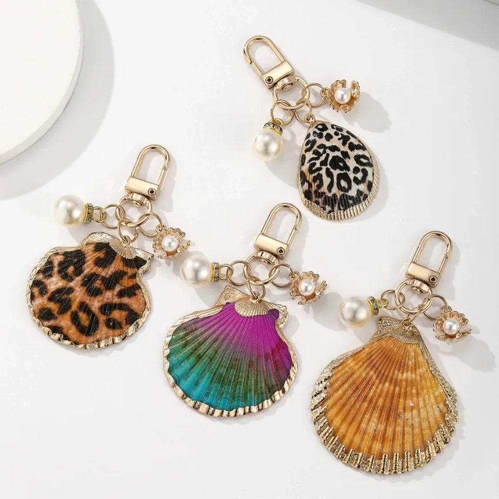 Four decorative shell keychain charms featuring leopard print, rainbow gradient, and natural orange shell designs with pearl beads and gold-tone metal hardware on a white background.