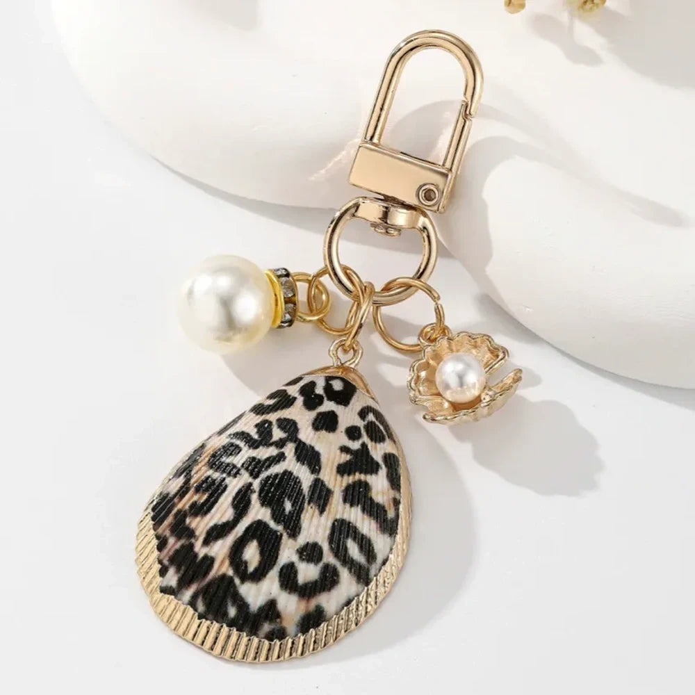 Four decorative shell keychain charms featuring leopard print, rainbow gradient, and natural orange shell designs with pearl beads and gold-tone metal hardware on a white background. - 2
