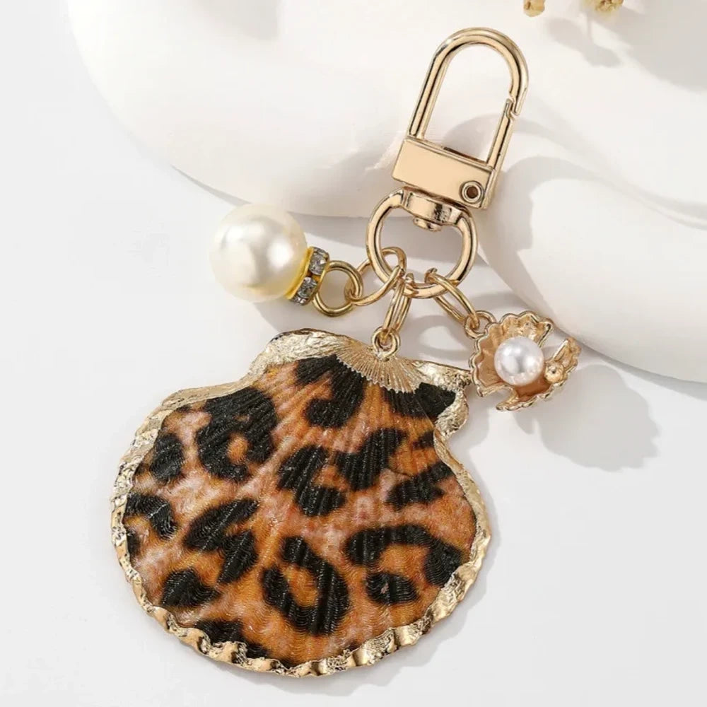 Four decorative shell keychain charms featuring leopard print, rainbow gradient, and natural orange shell designs with pearl beads and gold-tone metal hardware on a white background. - 4