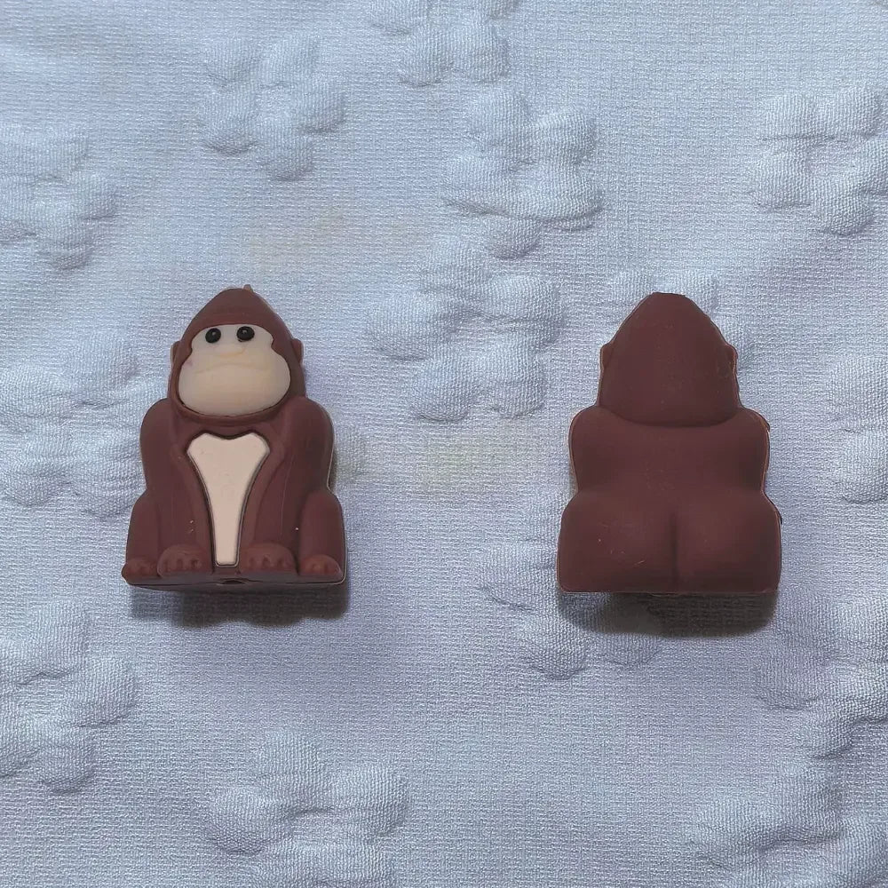 Nine 3D gorilla silicone focal beads arranged in a 3x3 grid, showing black and brown colorways of cartoon ape character beads on a white textured background. - Brown