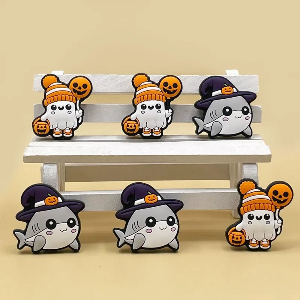 Cute Halloween silicone focal beads featuring ghost characters in orange knit hats with pumpkin balloons and shark characters in purple witch hats with jack-o-lantern accents, displayed on a miniature white bench.