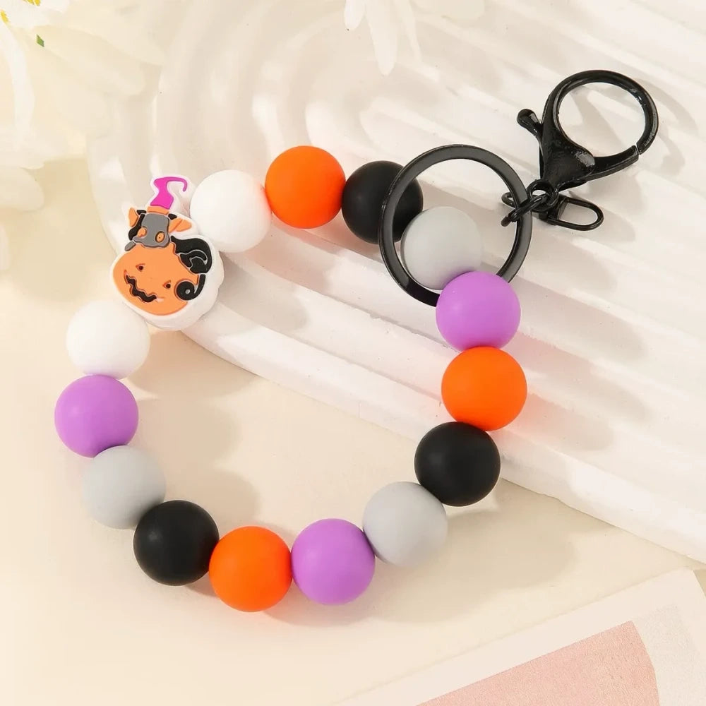Halloween themed printed silicone beads in orange, black, yellow, purple and white featuring pumpkin, skull, ghost and Happy Halloween designs, shown as completed keychain bracelets and phone charms. - 1