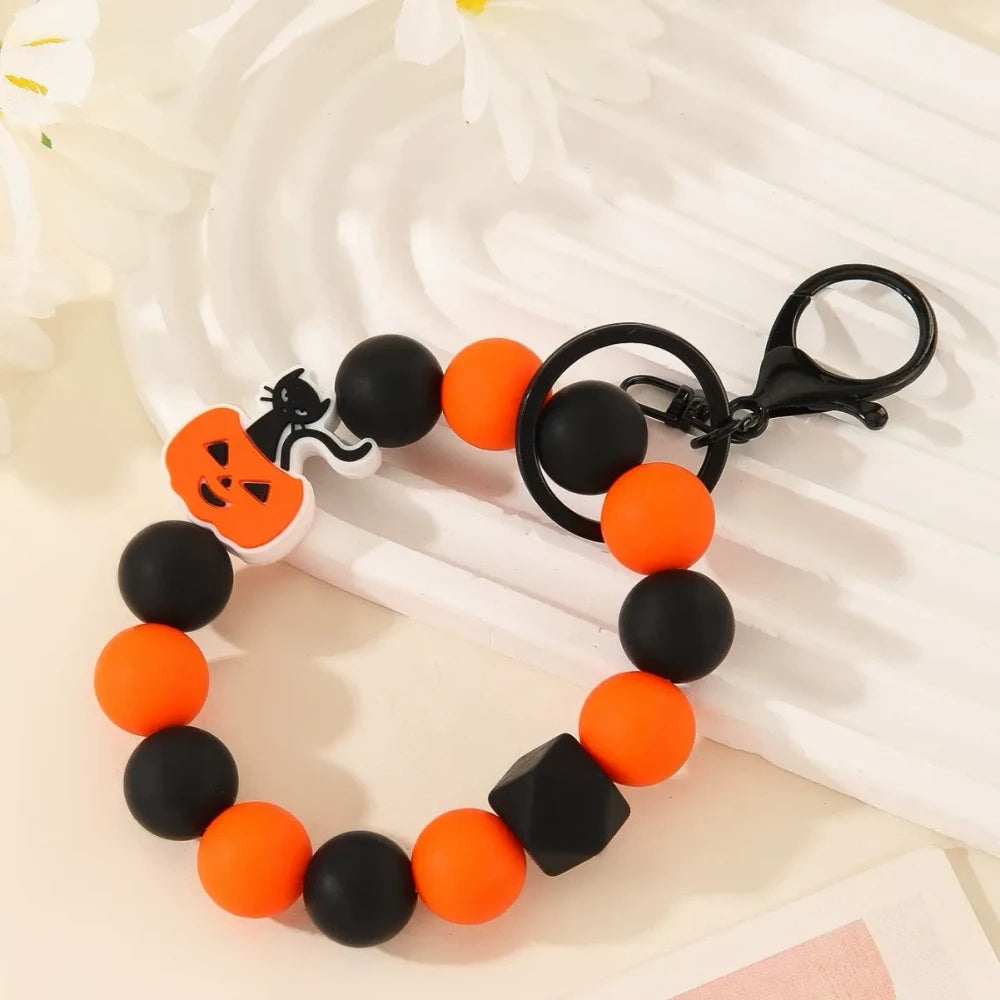 Halloween themed printed silicone beads in orange, black, yellow, purple and white featuring pumpkin, skull, ghost and Happy Halloween designs, shown as completed keychain bracelets and phone charms. - 4