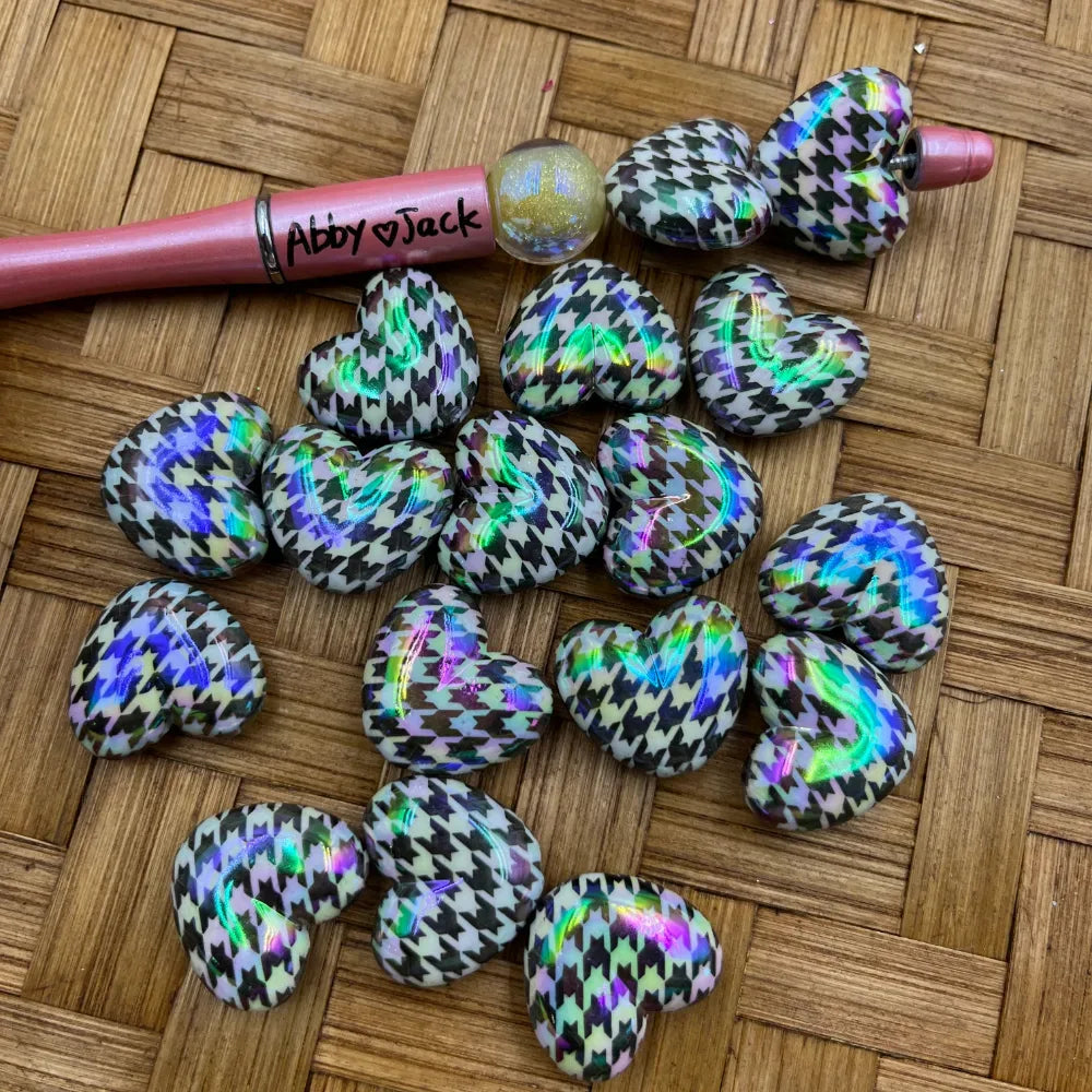 Holographic houndstooth print heart-shaped acrylic beads in black and white with iridescent rainbow shimmer, displayed on a woven surface next to a beadable pen.