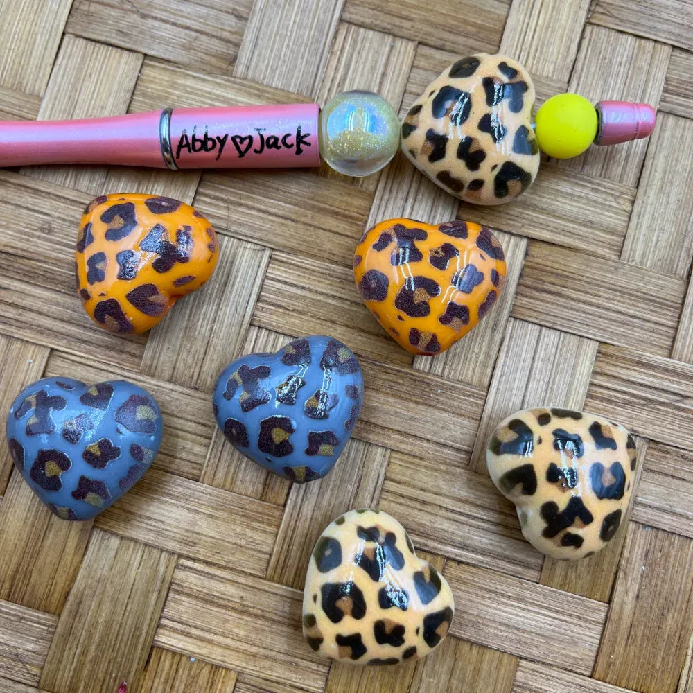 Assorted leopard print heart-shaped acrylic beads in orange, blue, and tan displayed on a woven surface next to a pink beadable pen labeled Abby Jack