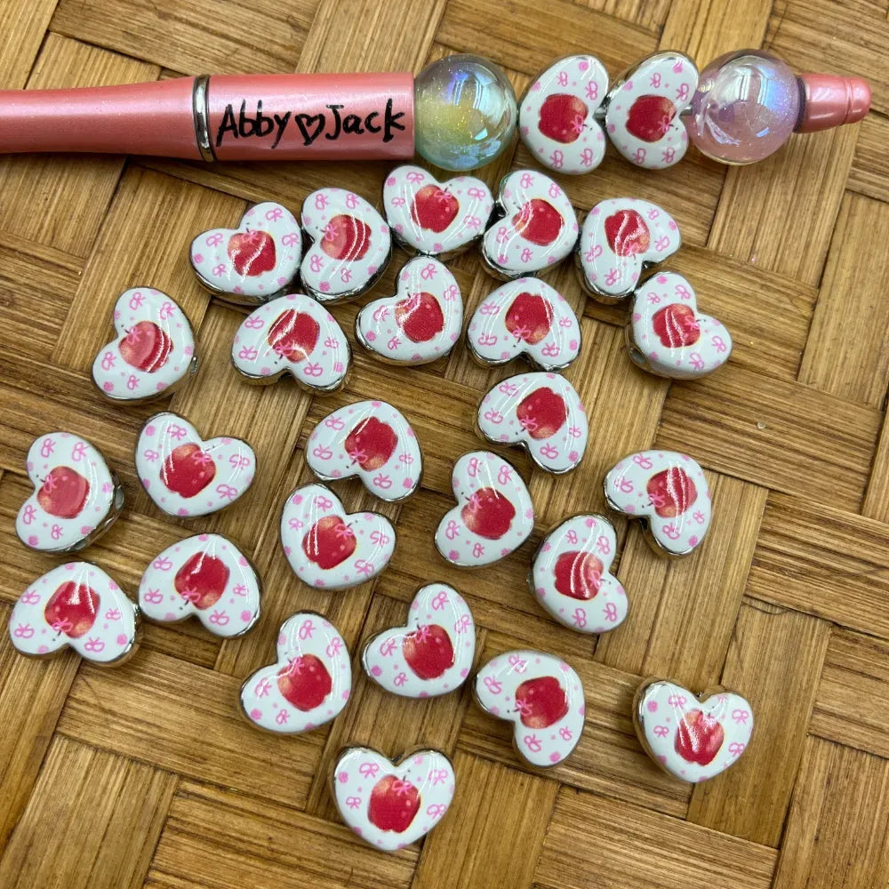 Heart-shaped white acrylic beads featuring red apple and pink bow pattern, shown with a pink beadable pen on wicker background
