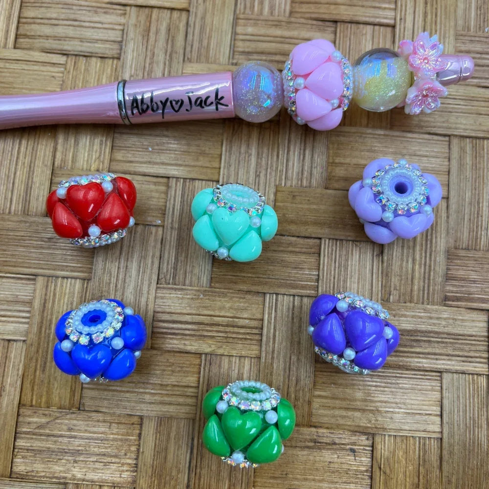 Colorful heart cluster fancy beads with rhinestone and pearl accents in red, teal, lavender, blue, purple, and green, displayed on woven surface next to a decorated pink beadable pen labeled Abby Jack.