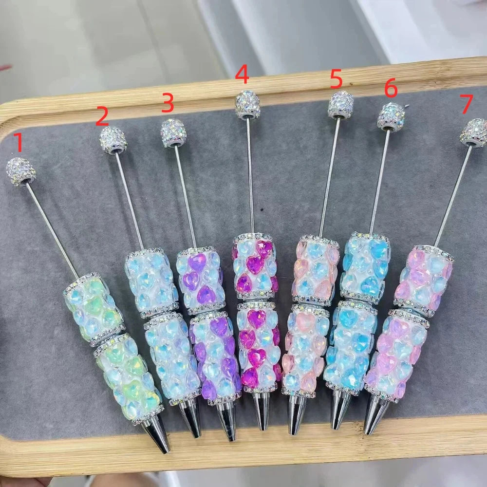 Seven rhinestone-encrusted beadable clay pens decorated with iridescent heart-shaped gems in pastel colors including mint green, blue, purple, pink and lavender, numbered 1 through 7, displayed on a wooden tray.