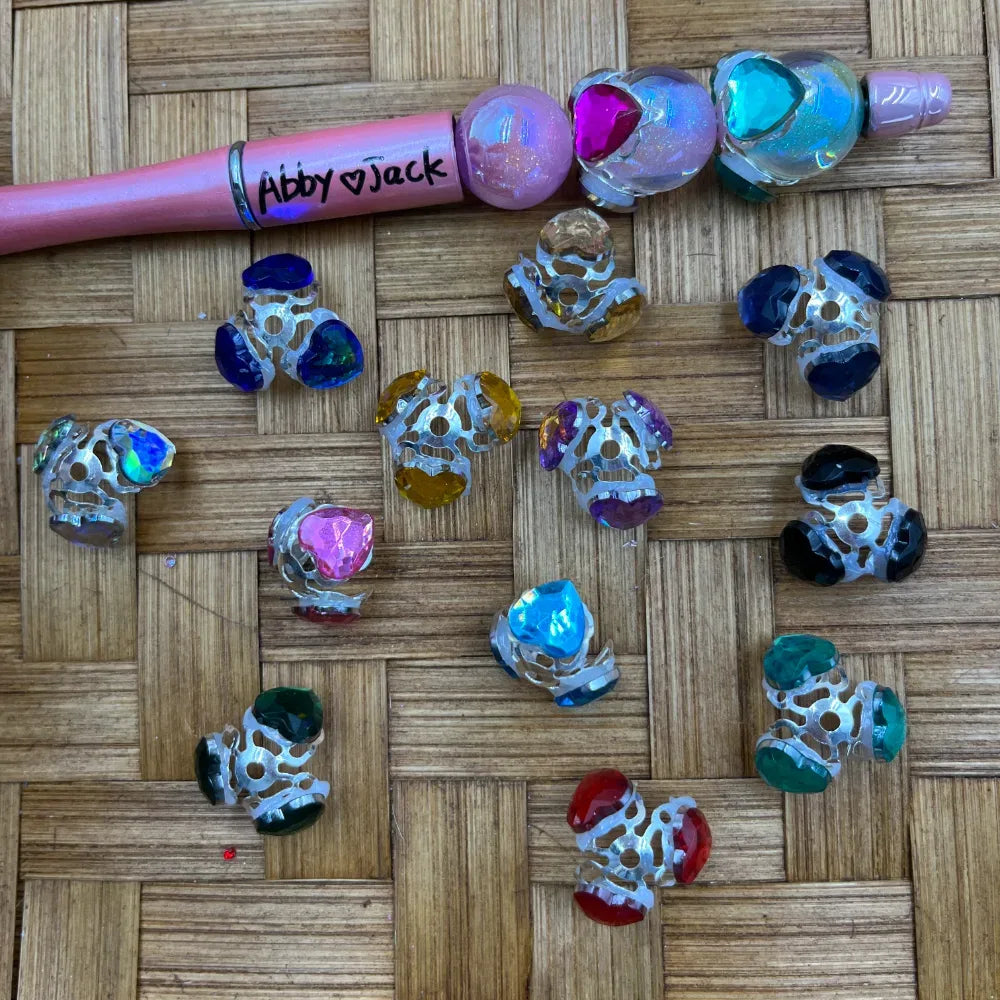 Assorted colorful heart gem silver filigree bead caps in multiple colors including blue, pink, teal, black, red, green, gold, and purple, displayed on a bamboo mat next to a pink beadable pen labeled Abby Jack.