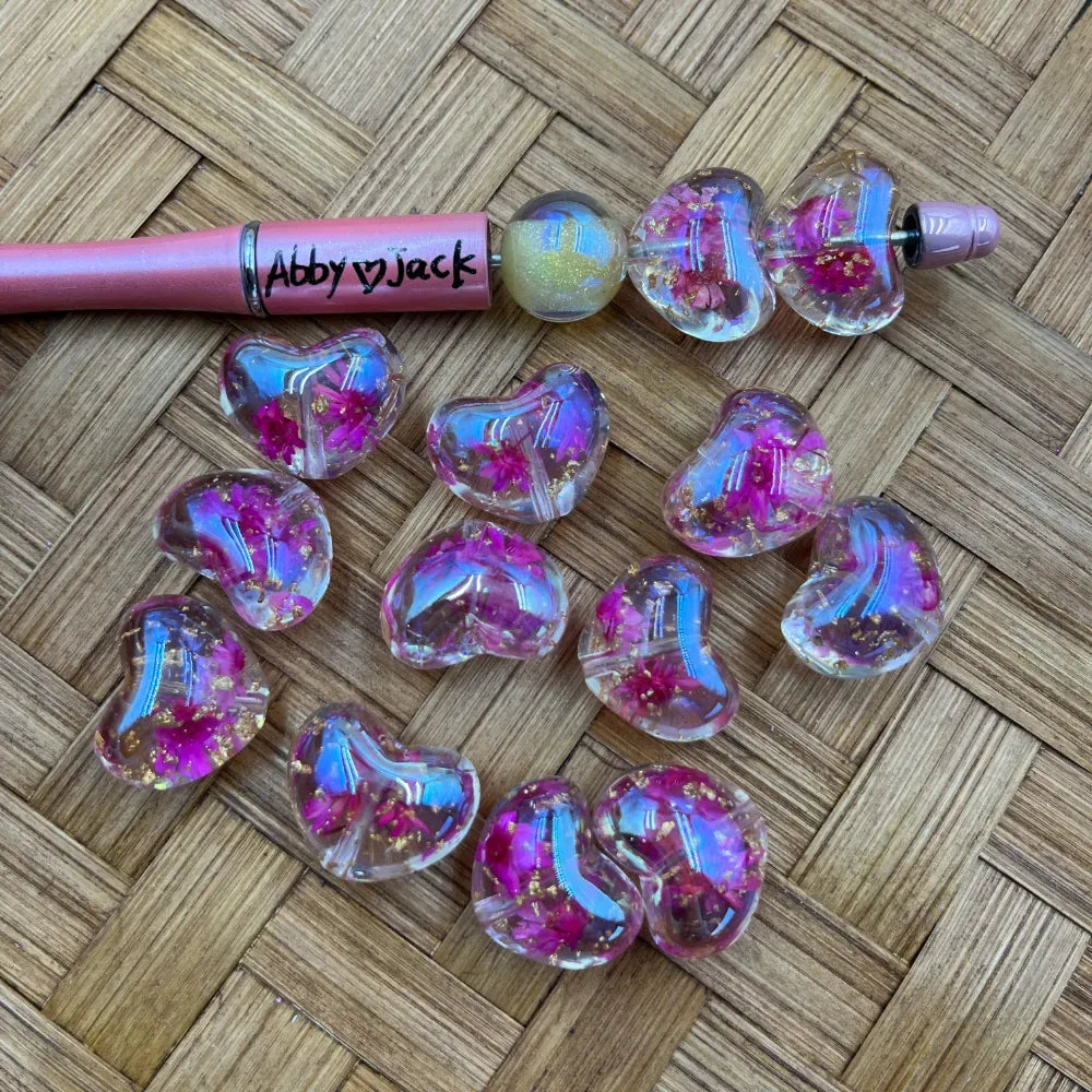 Clear iridescent heart-shaped acrylic beads with pink dried flowers and gold flakes, shown with a pink beadable pen on a woven bamboo surface