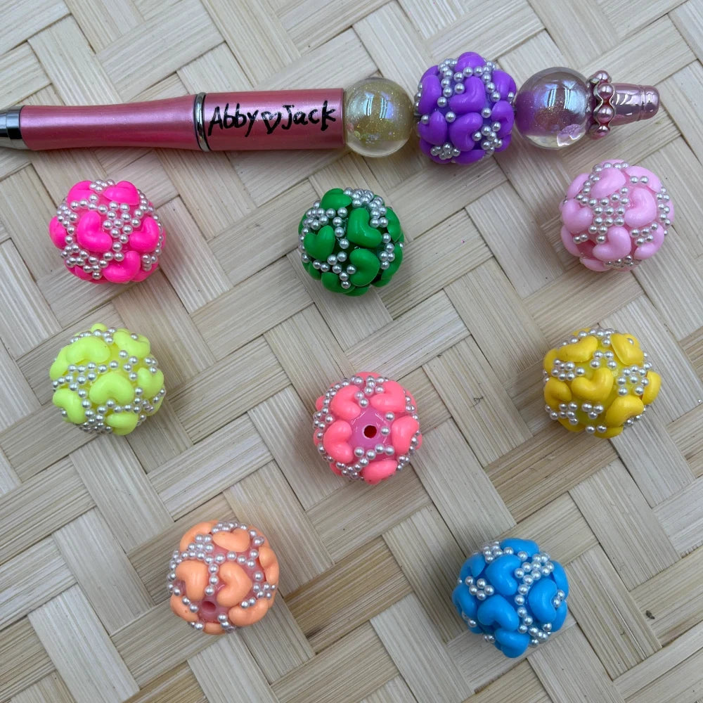 Colorful heart and pearl cluster chunky beads in pink, green, purple, yellow, orange, coral, blue, and lime arranged on woven bamboo surface with a decorated beadable pen.