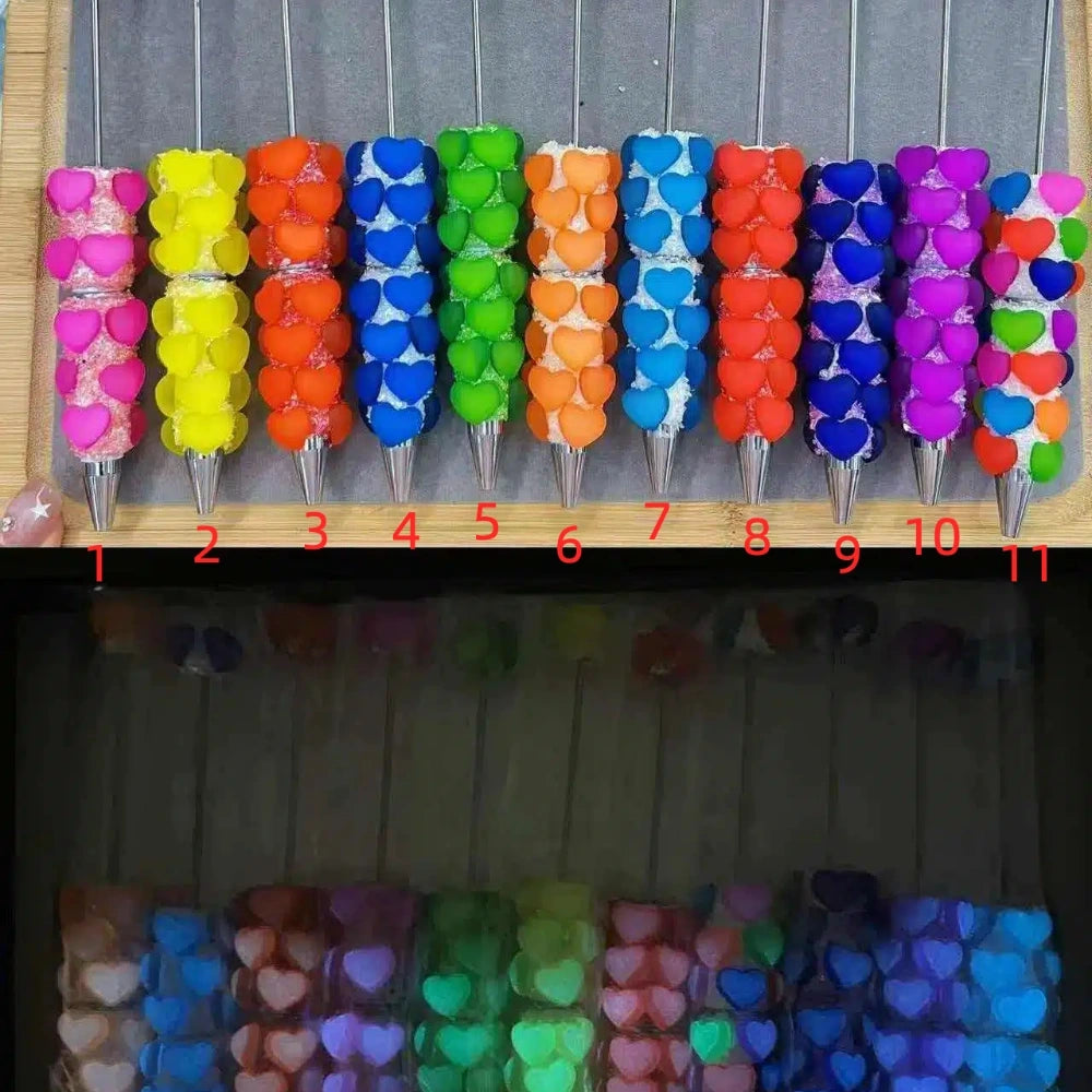 11 colorful heart rhinestone beadable pens arranged in a row including pink, yellow, orange, blue, green, purple and red varieties with silver tips on a wooden display board.