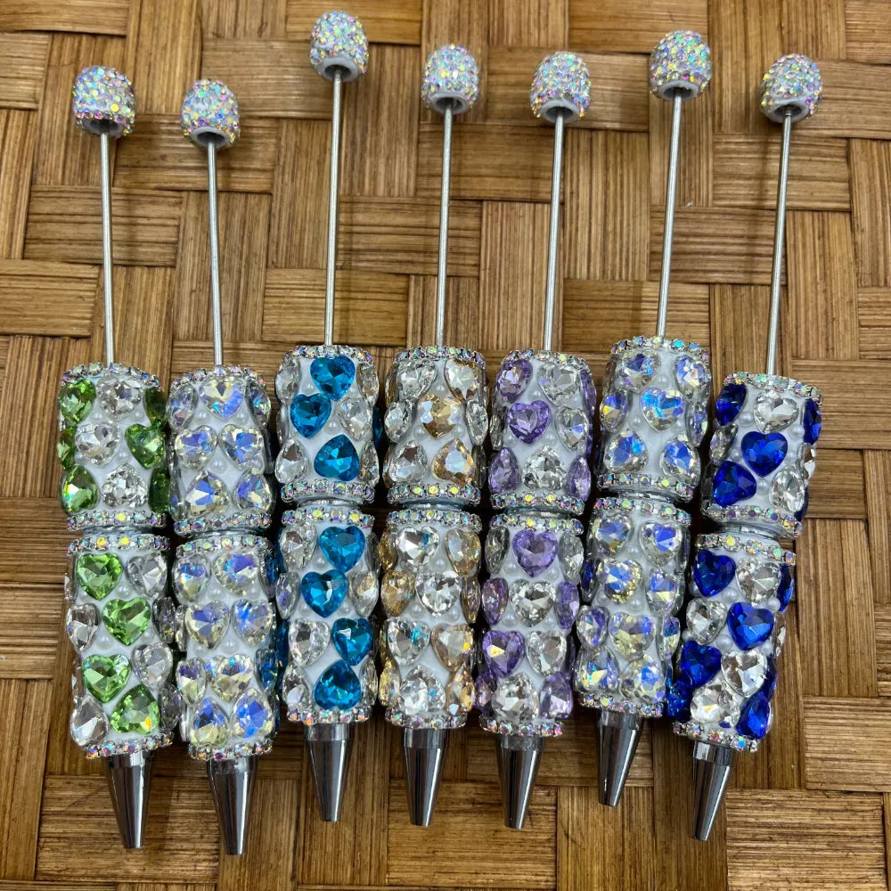 Five sparkling heart-shaped rhinestone crystal clay beadable pens in green, teal, champagne, purple, and blue colors with glitter ball toppers on a woven bamboo background.