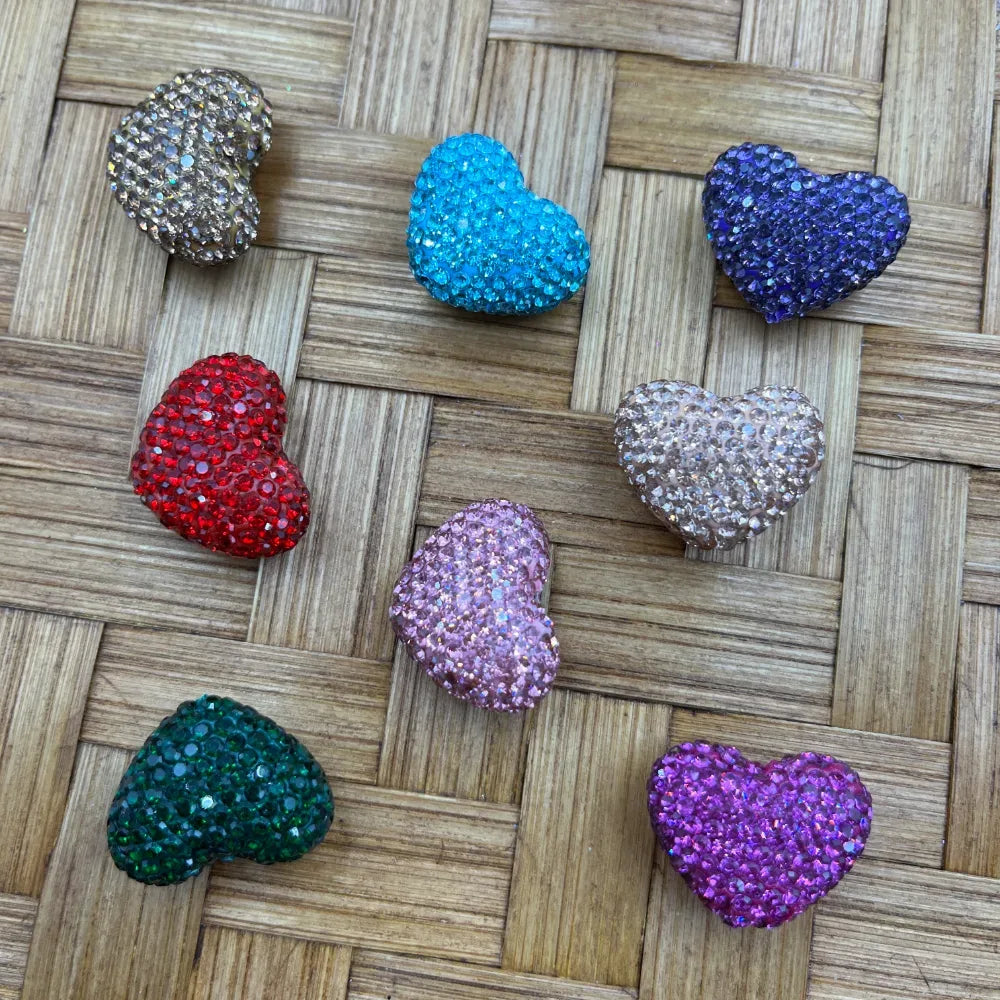 Eight rhinestone-covered heart shaped beads in assorted colors including red, blue, purple, teal, pink, lavender, champagne, and silver on a woven bamboo surface.