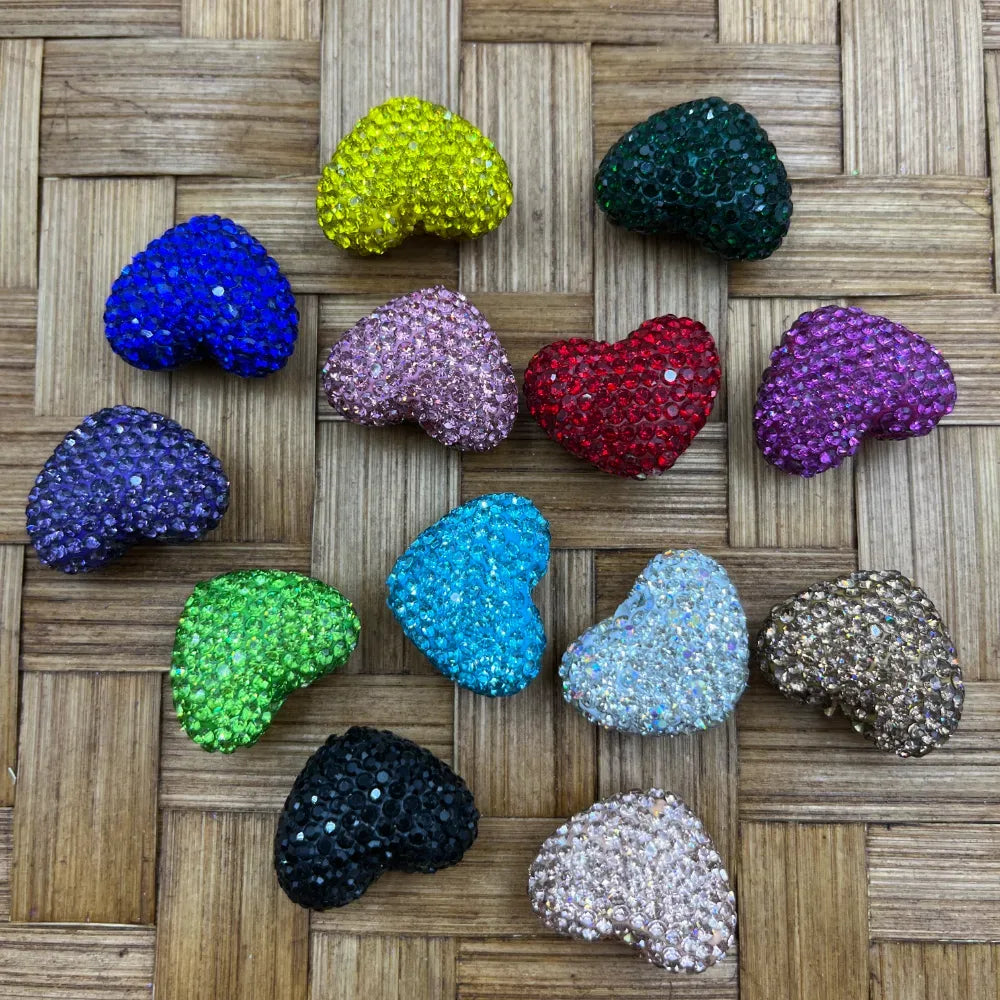 Assorted colorful heart-shaped rhinestone beads in multiple colors including red, blue, green, pink, purple, black, teal, gold, and clear on a woven bamboo surface.