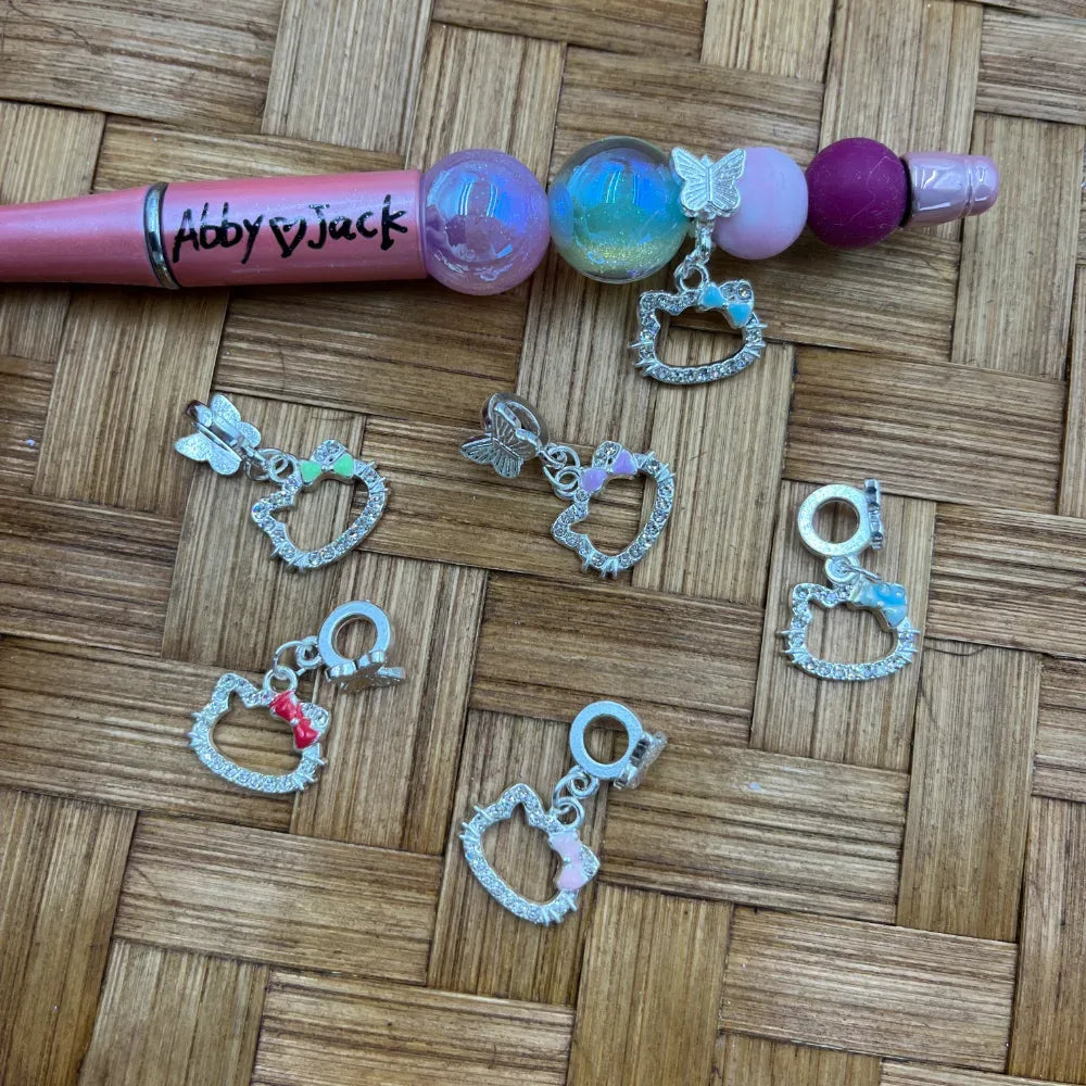Assorted Hello Kitty shaped silver rhinestone bail beads with colorful bow accents and butterfly charms displayed on a woven surface next to a beadable pen