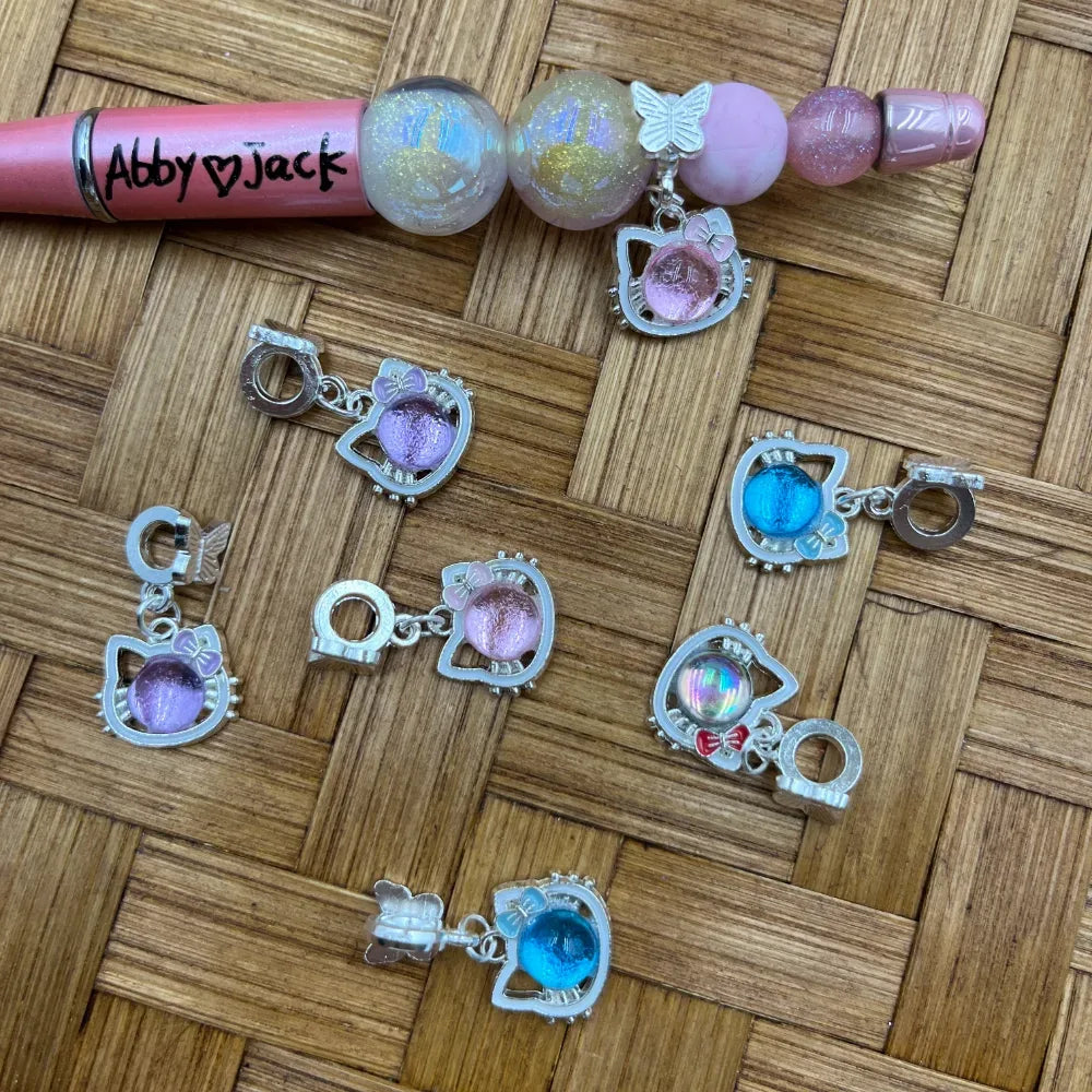 Hello Kitty shaped silver bail beads with colorful iridescent and glitter ball charms in purple, pink, blue, and holographic, displayed on a wicker surface alongside a decorated beadable pen.