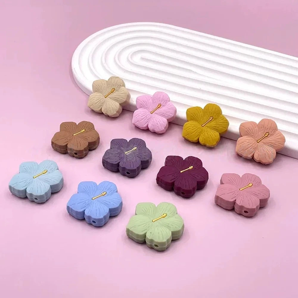 Assorted pastel and earthy tone hibiscus flower silicone focal beads with gold stamen detail displayed on a pink surface, available in colors including blue, purple, green, pink, mauve, brown, tan, yellow, and peach.