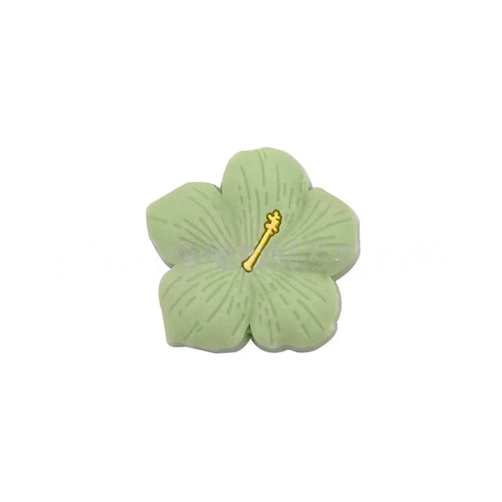 Assorted pastel and earthy tone hibiscus flower silicone focal beads with gold stamen detail displayed on a pink surface, available in colors including blue, purple, green, pink, mauve, brown, tan, yellow, and peach. - Matcha Green