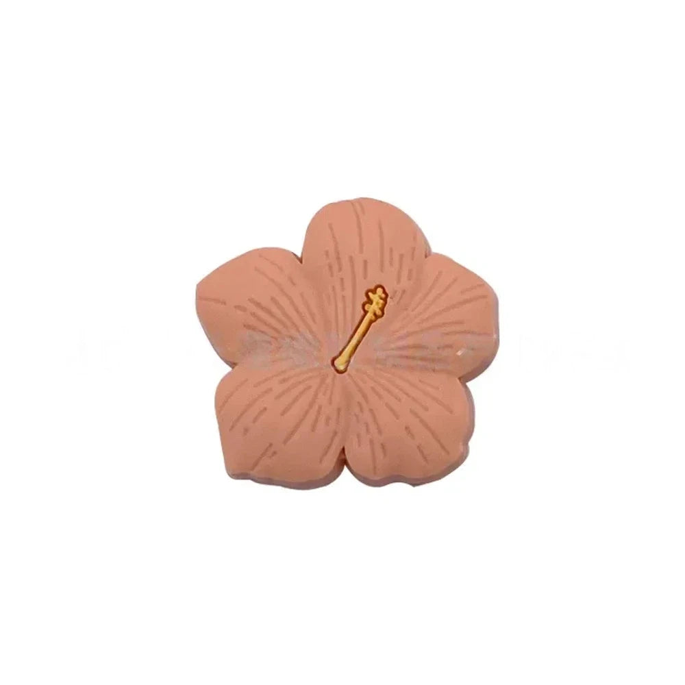 Assorted pastel and earthy tone hibiscus flower silicone focal beads with gold stamen detail displayed on a pink surface, available in colors including blue, purple, green, pink, mauve, brown, tan, yellow, and peach. - Orange