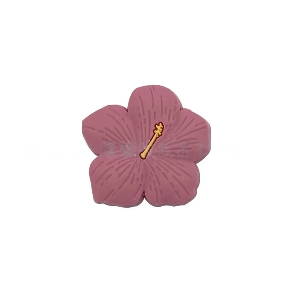 Assorted pastel and earthy tone hibiscus flower silicone focal beads with gold stamen detail displayed on a pink surface, available in colors including blue, purple, green, pink, mauve, brown, tan, yellow, and peach. - Pink