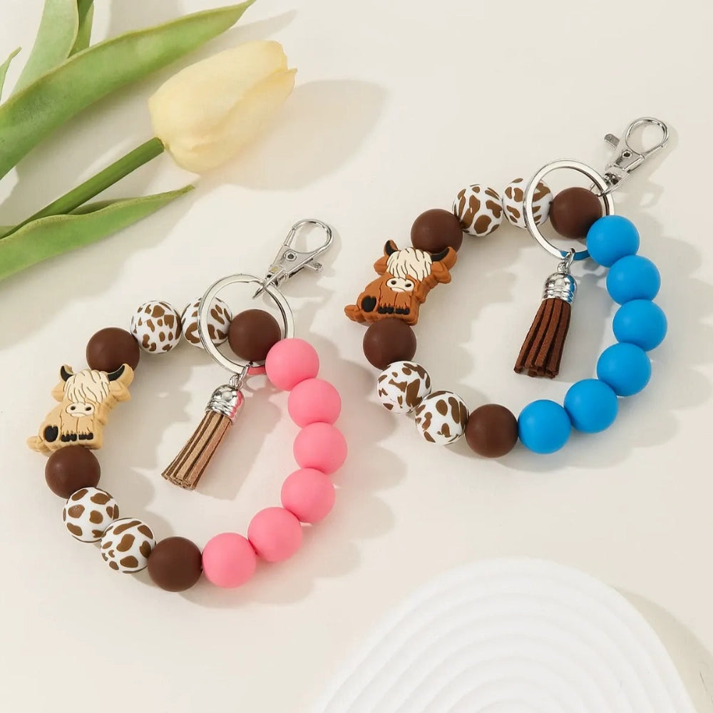 Silicone bead bracelet keychains featuring highland cow charms, cow print beads, and solid pink and blue silicone beads with tassel accents.