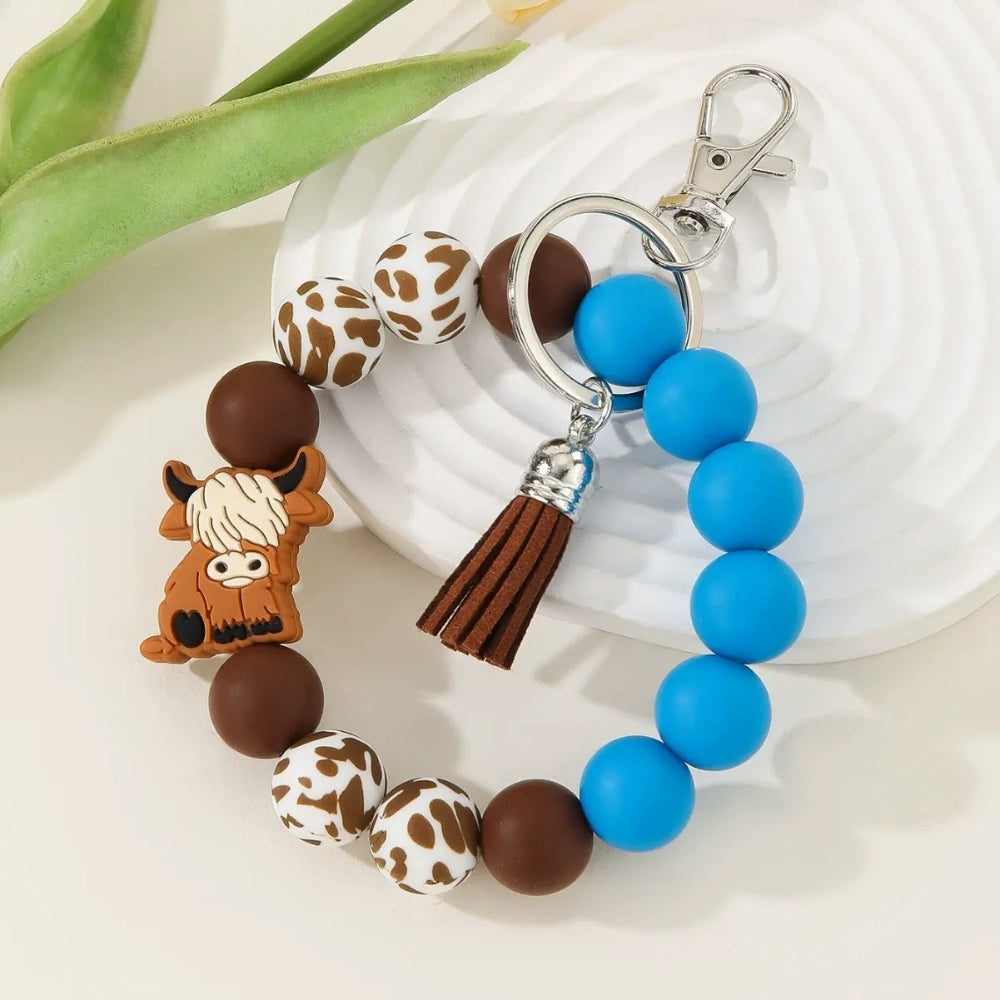 Silicone bead bracelet keychains featuring highland cow charms, cow print beads, and solid pink and blue silicone beads with tassel accents. - Pink
