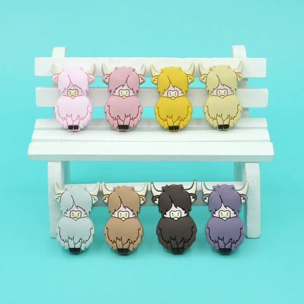 Eight colorful silicone highland cow focal beads in pink, mauve, yellow, cream, grey, brown, dark brown, and purple displayed on a miniature white wooden bench against a teal background. - Random Mixed