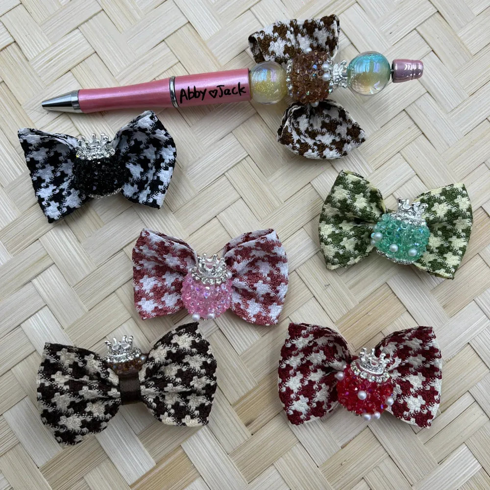 Six houndstooth fabric bow embellishments in black, brown, pink, green, dark brown, and red with silver crown and bead center decorations, displayed with a pink beadable pen on woven background.