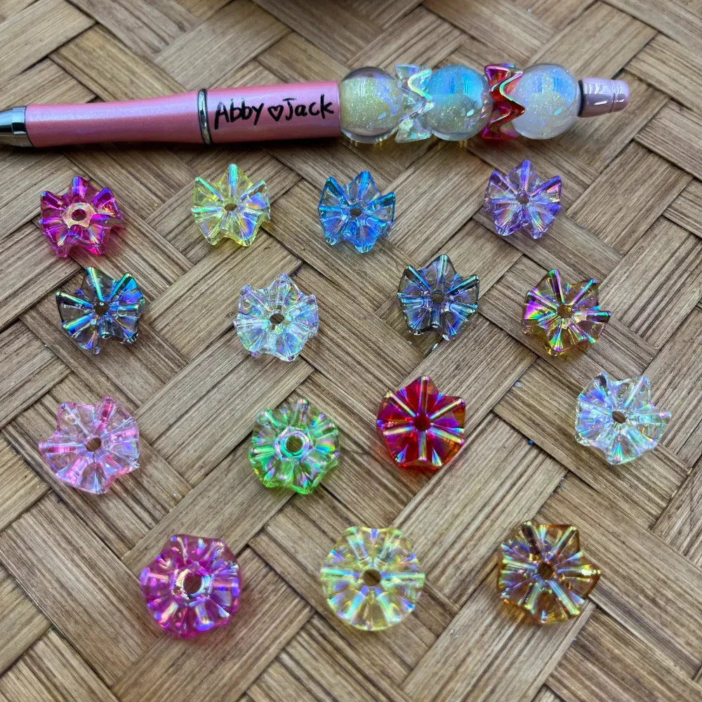 Assorted iridescent AB bow-shaped acrylic spacer beads in pink, blue, green, red, yellow, and clear displayed on wicker surface next to a beadable pen labeled Abby Jack.