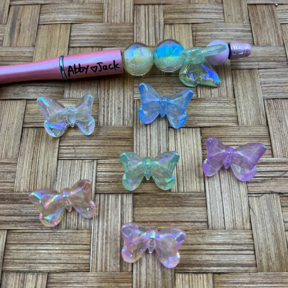 Iridescent AB pastel butterfly acrylic beads in clear, blue, green, pink, and purple displayed on a woven surface next to a pink beadable pen labeled Abby Jack.