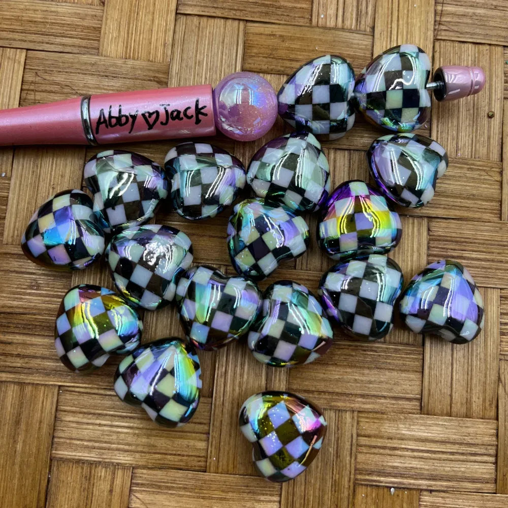 Iridescent black and white checkered heart-shaped acrylic beads with AB rainbow finish displayed on a woven surface next to a beadable pen labeled Abby Jack.