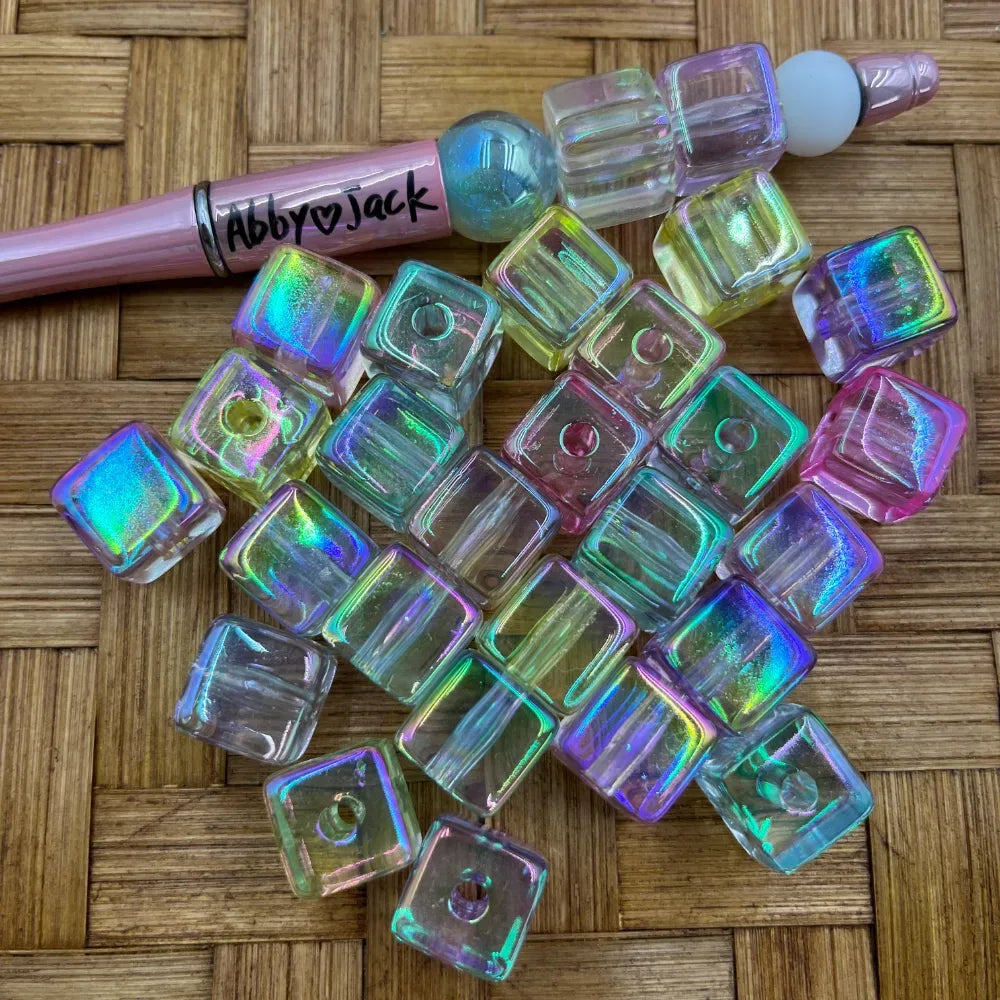 Iridescent holographic AB clear cube acrylic beads scattered on wicker surface next to a pink beadable pen branded Abby Jack, showing rainbow color-shifting effect.