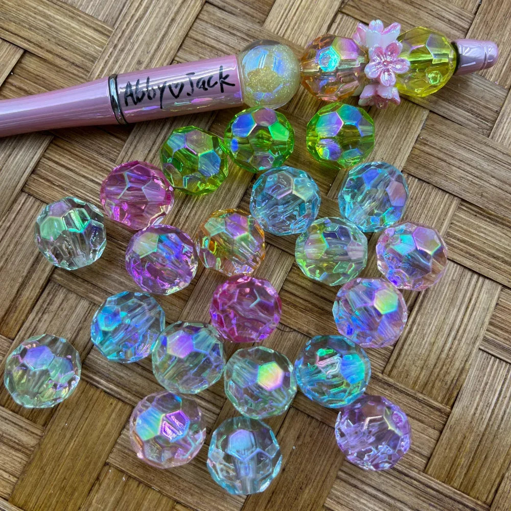 Iridescent AB faceted round acrylic beads in mixed pastel colors including pink, green, blue, and clear displayed on a woven surface next to a decorated beadable pen.