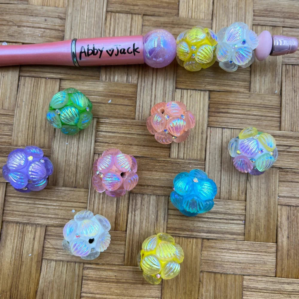 Iridescent AB flower cluster acrylic fancy beads in assorted pastel colors including purple, pink, green, blue, yellow, and white with rhinestone accents, displayed on woven bamboo surface next to a beadable pen labeled Abby and Jack.
