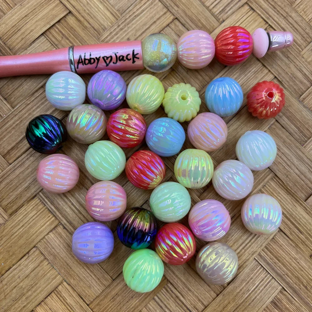 Assorted iridescent AB melon ribbed acrylic beads in pastel pink, purple, yellow, green, red, and dark multicolor finishes displayed on a woven surface next to a pink beadable pen labeled Abby and Jack.