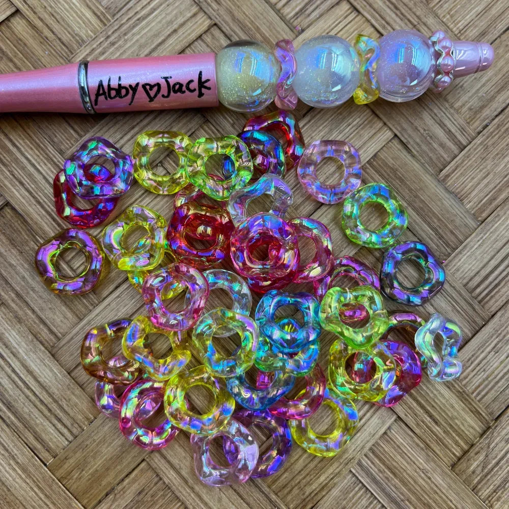 Colorful iridescent AB rainbow wavy ring acrylic spacer beads in mixed colors including pink, purple, yellow, green, and clear, scattered on a woven surface next to a pink beadable pen decorated with globe beads.