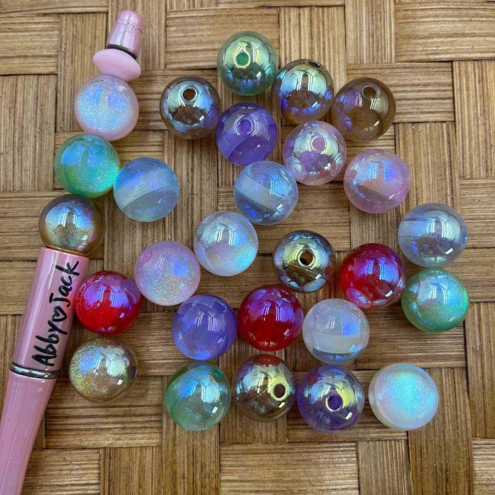 Mixed iridescent aurora AB acrylic beads in pastel purple, red, clear, green and blue with holographic shimmer finish displayed on woven surface next to a pink beadable pen