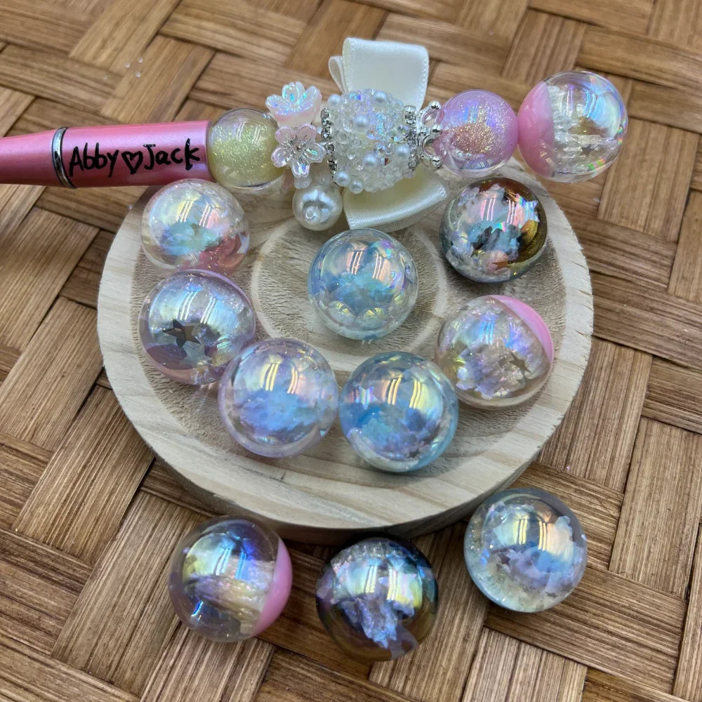 Iridescent holographic aurora bubble ball pen toppers with glitter and star confetti fill displayed on a wooden disc, featuring a pink beadable pen for scale