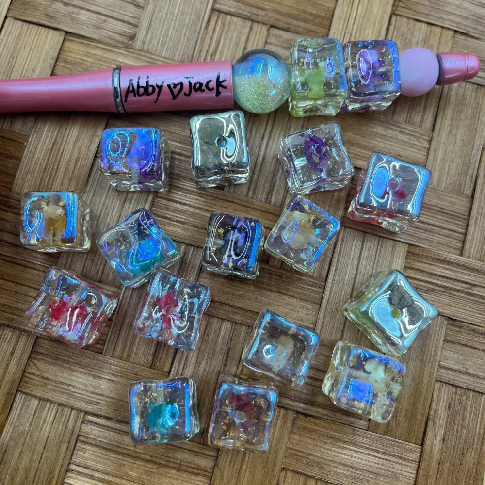 Assorted iridescent aurora square cube acrylic beads in mixed colors with holographic shimmer effect displayed next to a pink beadable pen on a woven surface