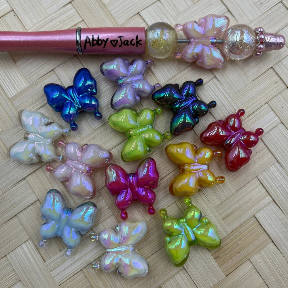 Assorted iridescent pearlescent butterfly acrylic beads in 11 colors including blue, purple, pink, white, green, red, gold, and magenta, displayed next to a pink beadable pen on a wicker surface.