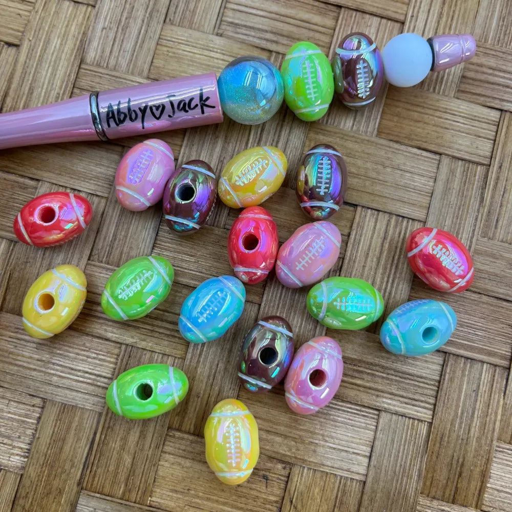 Assorted iridescent football-shaped acrylic beads in multiple colors including red, green, yellow, pink, blue, and brown, displayed on a wicker surface next to a pink beadable pen labeled Abby Jack.