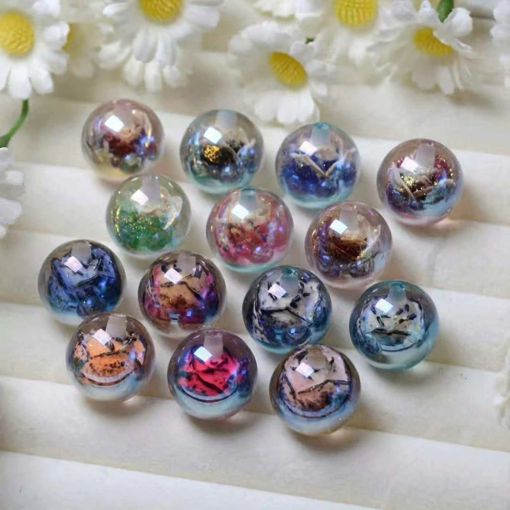 13 round iridescent acrylic beads with colorful dried flower and glitter galaxy inclusions in purple, blue, pink, and green tones arranged on white fabric with daisies in background